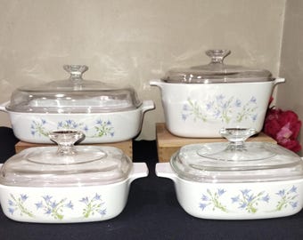 May include: Set of four white casserole dishes with glass lids. Each dish features a floral design with blue flowers and green leaves.