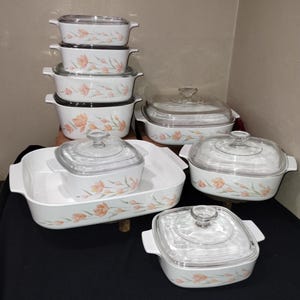 May include: A collection of white ceramic baking dishes with clear glass lids. The dishes feature a floral pattern in shades of peach and green. Various sizes are stacked and arranged, showcasing a complete bakeware set.