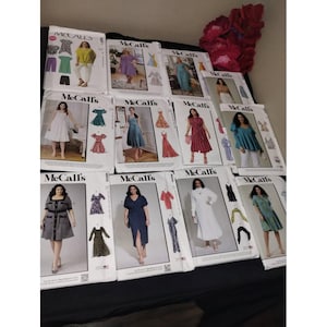 May include: A collection of McCall's sewing pattern envelopes. The envelopes feature images of dresses, tops, and pants. The patterns are displayed on a black surface with a cluster of red flowers in the background.