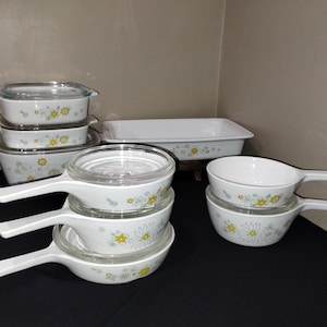 May include: A set of vintage Pyrex baking dishes with a floral pattern in shades of yellow, green, and blue. The set includes a variety of sizes and shapes, including a rectangular baking dish, round casserole dishes, and a small saucepan.