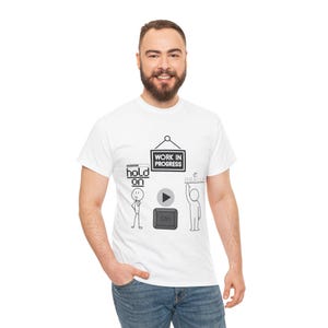 May include: A white short-sleeved t-shirt featuring a black graphic print. The design includes a sign reading WORK IN PROGRESS, a stick figure with the text hold on and a loading bar, a play button, a Ctrl key icon, and a stick figure with the text REALLY.