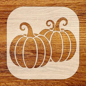Könnte beinhalten: Eine Holzschablone mit zwei Kürbisumrissen. Die Kürbisse haben gebogene Stiele und vertikale Linien, um die Rippen des Kürbisses darzustellen. Die Schablone befindet sich vor einem hellen Holzmaserungshintergrund.