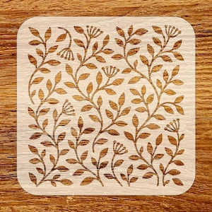 May include: Square wooden coaster with a laser-cut floral design. The coaster features a light wood background with a repeating pattern of brown leaves and flowers. The edges of the coaster are rounded.
