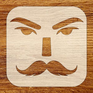 May include: A square stencil featuring a stylized face with a mustache, nose, eyebrows, and eyes. The face is cut out of a light-colored material, set against a wooden background. The mustache has curved ends, and the eyebrows are arched.
