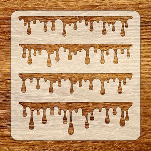 Puede incluir: Un esténcil de madera con cuatro filas horizontales de diseños de pintura goteando. La pintura goteando es de color marrón medio, y el esténcil en sí es de color beige claro. El esténcil es cuadrado con esquinas redondeadas.