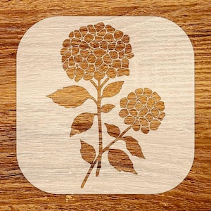 May include: A wooden coaster featuring a laser-cut hydrangea design. The flower and leaves are a darker brown against a light wood background. The coaster has rounded corners.