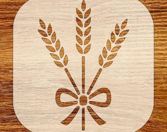 May include: A wheat stalk stencil with three wheat bundles tied with a bow. The stencil is light beige with a rounded square shape. The wheat and bow are a darker brown color. The background is a wooden surface.