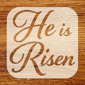 Pode incluir: Um estêncil de madeira com as palavras "He is Risen" recortadas em uma escrita fluida. O estêncil é de cor clara com formato quadrado e cantos arredondados. O texto é em uma cor marrom quente.