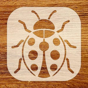 Puede incluir: Un esténcil de mariquita blanco con esquinas redondeadas, con un diseño detallado de insecto. La mariquita tiene un cuerpo segmentado con puntos y antenas, perfecto para proyectos de manualidades. El esténcil está sobre un fondo de madera.