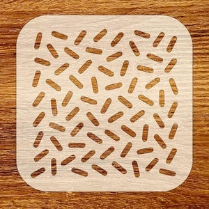 May include: A square, light-colored stencil with rounded corners. The stencil features a repeating pattern of small, oblong cutouts. The background is a wooden surface with visible grain.