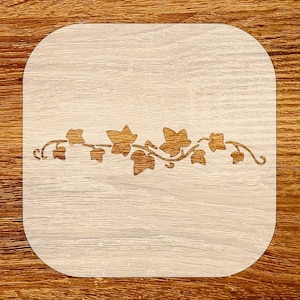 May include: A light-colored wooden coaster with rounded corners. A decorative vine design with leaves is etched across the center. The coaster is set against a wooden surface.