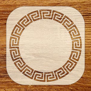 Puede incluir: Un posavasos cuadrado de madera clara con un diseño de clave griega tallada y repetida alrededor de un círculo central. El posavasos se encuentra sobre una superficie de madera más oscura, creando un contraste de tonos. El diseño es un elemento decorativo clásico.