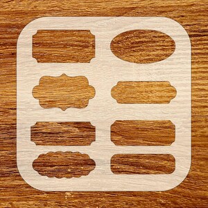 May include: A white stencil with various label shapes, including rectangles, ovals, and decorative frames. The stencil is placed on a wooden surface, showcasing the design's potential for crafting and DIY projects.