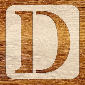 May include: A square stencil featuring a large, bold, serif-style capital letter D cut out from the center. The stencil material is a light, neutral-toned sheet with a smooth surface, contrasting against the dark, horizontal wood grain pattern.