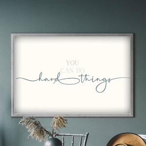 Könnte beinhalten: Ein weißer gerahmter Druck mit dem Text "You can do hard things" in blauer und grauer Schrift auf weißem Hintergrund. Der Druck hängt an einer blaugrünen Wand über einem Holztisch mit Stühlen.