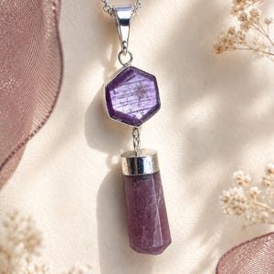 May include: A silver necklace featuring a hexagonal purple gemstone pendant connected to a cylindrical purple crystal drop. The pendant is set in polished silver metal and hangs from a delicate silver chain, showcasing natural stone textures.