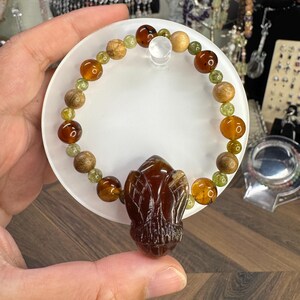 Puede incluir: Una pulsera de cuentas con un colgante marrón tallado. La pulsera incluye cuentas de color ámbar, verde y madera. La pulsera se muestra sobre una superficie circular blanca.