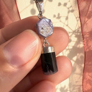 May include: A pendant necklace featuring a hexagonal, light purple, translucent gemstone set in silver, connected to a cylindrical, opaque black stone drop. The pendant hangs from a silver-toned metal chain, showcasing a dual-stone design.