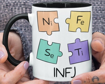 May include: White ceramic mug with a black rim and handle. The mug has a design of four puzzle pieces with the letters "Ni", "Fe", "Se", and "Ti" on them. The text "INFJ" is printed below the puzzle pieces.