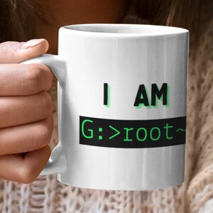 Könnte beinhalten: Weiße Keramiktasse mit dem Text "I AM G:>root~" in Schwarz und Grün. Die Tasse wird von einer Person gehalten. Die Tasse hat eine Standardgröße und -form, geeignet für Kaffee, Tee oder andere Getränke. Ein tolles Geschenk für Programmierer.