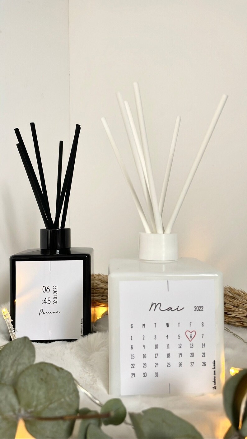 May include: Two glass diffuser bottles with black and white reeds. The black bottle has a label with the text "06:45 02.01.2022 Perine". The white bottle has a label with the text "Mai 2022" and a calendar with a heart on the 6th.