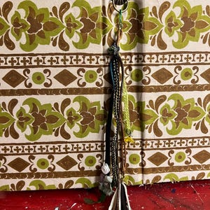 May include: A decorative tassel with various textures and colors, including black, brown, and cream, hangs from a metal ring. The background features a patterned fabric with green and brown floral and geometric designs.