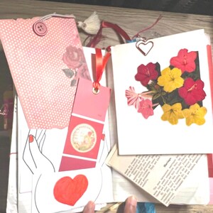 May include: A collection of handmade paper crafts, including a pink polka dot gift bag with a button closure, a red bookmark, and a card with pressed flowers. A heart-shaped paper clip and a drawing of hands holding a heart are also visible.