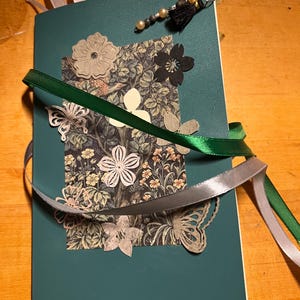 May include: A teal green journal with a floral design on the front cover. The journal is decorated with ribbons, a butterfly, and a charm with beads and a tassel.