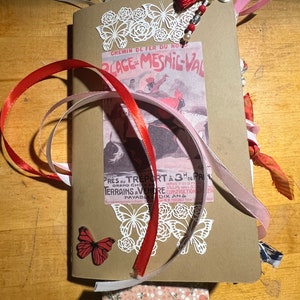 May include: A brown paper journal with a vintage poster of a horse race on the front. The journal is decorated with red and pink ribbons, lace, and paper butterflies. The poster text reads "Chemin de Fer du Nord Place de Mesnil-Val".