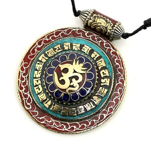 May include: A round pendant necklace with a gold Om symbol surrounded by a blue and turquoise mandala. The pendant is framed by a red and gold border with intricate designs. The necklace is made of black cord.