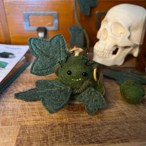 May include: A handmade felt creature with a whimsical design. The green figure has large, leaf-shaped wings and a mushroom on its head. The creature has small black eyes and a smiling mouth. A small red ladybird sits on top of the mushroom.