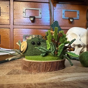 May include: A felted sculpture of a green creature with button eyes and a wooden button detail, set on a wood slice base. The sculpture is surrounded by felted greenery and brown bead accents, creating a miniature forest scene.