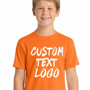 May include: An orange t-shirt with the words "CUSTOM TEXT LOGO" in white, displayed on the front. The shirt is short-sleeved and worn by a person. The person is wearing blue jeans.