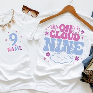May include: Two white t-shirts. The left shirt features the number 9 and the word NAME in blue and pink. The right shirt displays the text ON CLOUD NINE in blue and pink, accompanied by a smiley face, a rainbow, stars, flowers, and a cloud.