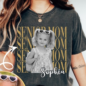Pu&ograve; includere: Una t-shirt grigio antracite con il testo dorato ripetuto SENIOR MOM e una fotografia in bianco e nero di una bambina con le codine che indossa occhiali da sole a forma di cuore e uno zaino, con il nome Sophia scritto in bianco sotto.
