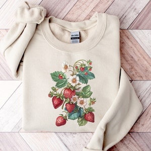 Pode incluir: Uma camisola de cor creme com uma ilustra&ccedil;&atilde;o vintage de morangos e flores brancas. O design est&aacute; centrado na frente da camisola.