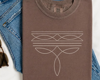 May include: A brown short-sleeve t-shirt featuring a white line art graphic of geometric shapes and curved lines on the chest. The shirt has a crew neckline and a Comfort Colors label at the inner collar. Blue denim jeans and round sunglasses.