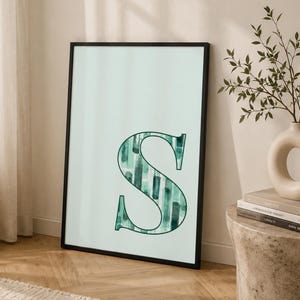 May include: A rectangular wall art print in a thin black frame featuring a large capital letter S centered on a pale mint background. The letter S is filled with a vertical pattern of dark green and teal watercolor-style brush strokes.