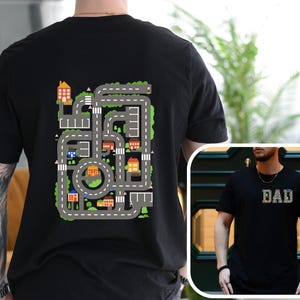 May include: Black t-shirt featuring a colorful road map design on the back with houses, cars, and trees. The shirt also has the word "DAD" on the front. The shirt is made of a soft material.