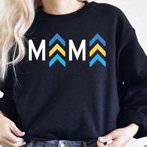 May include: Black sweatshirt with the word "MAMA" in white letters and blue and yellow chevron arrows.