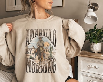 May include: A beige sweatshirt with a vintage graphic print of a cowboy riding a horse in a desert landscape. The graphic text reads "Amarillo By Morning".