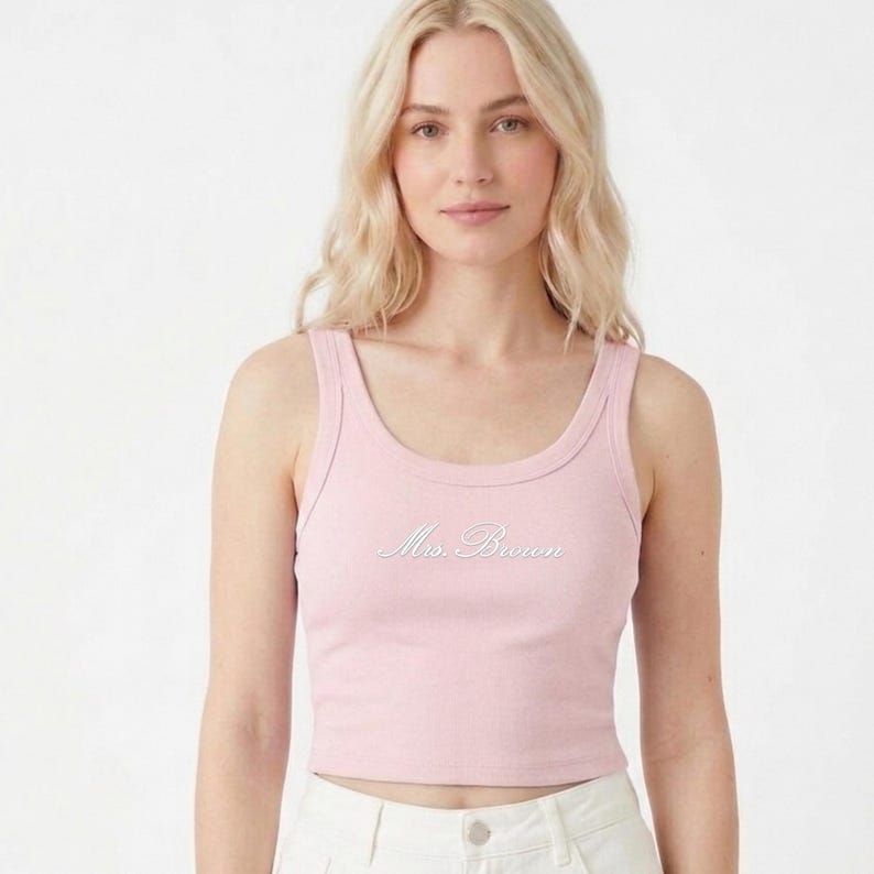 May include: A light pink, scoop-neck crop top with the cursive text "Ms. Brown" embroidered on the front. The top is sleeveless and made of a soft, ribbed material. The model is wearing white pants.