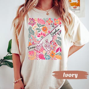 Op de afbeelding: Ivoor kleurig T-shirt met een kleurrijk bloemenontwerp met roze, oranje en blauwe bloemen. De afbeelding toont twee handen die elkaar vasthouden, met de namen "Emma" en "Sophia" erboven. Het shirt heeft een casual stijl.