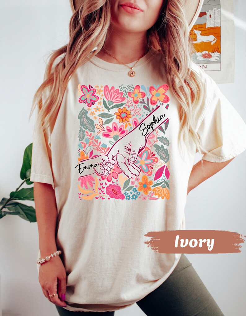 Op de afbeelding: Ivoor kleurig T-shirt met een kleurrijk bloemenontwerp met roze, oranje en blauwe bloemen. De afbeelding toont twee handen die elkaar vasthouden, met de namen "Emma" en "Sophia" erboven. Het shirt heeft een casual stijl.