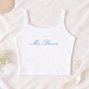 May include: White ribbed tank top with thin straps. The text "Mrs. Brown" is embroidered in light blue script across the front. The top is a simple, elegant design, perfect for casual wear or layering.