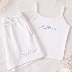 May include: White pajama set featuring a camisole top and shorts. The top has thin straps and the embroidered text "Mrs. Brown" in blue. The shorts have an elastic waistband and the embroidered date "06.11.2026" in blue. The set is made of a soft material.