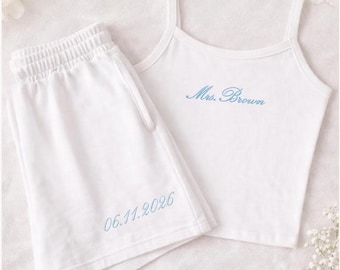 May include: White pajama set featuring a camisole top and shorts. The top has thin straps and the embroidered text "Mrs. Brown" in blue. The shorts have an elastic waistband and the embroidered date "06.11.2026" in blue.