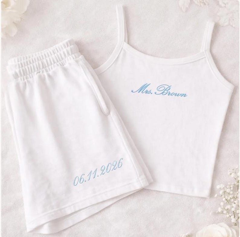 May include: White pajama set featuring a camisole top and shorts. The top has thin straps and the embroidered text "Mrs. Brown" in blue. The shorts have an elastic waistband and the embroidered date "06.11.2026" in blue. The set is made of a soft material.