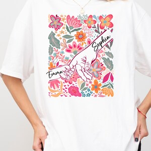 Op de afbeelding: Wit T-shirt met een bloemenontwerp met roze, oranje en blauwe bloemen. De afbeelding toont twee handen die elkaar vasthouden, met de namen "Emma" en "Sophia" erop. Een casual kledingstuk voor dagelijks gebruik.