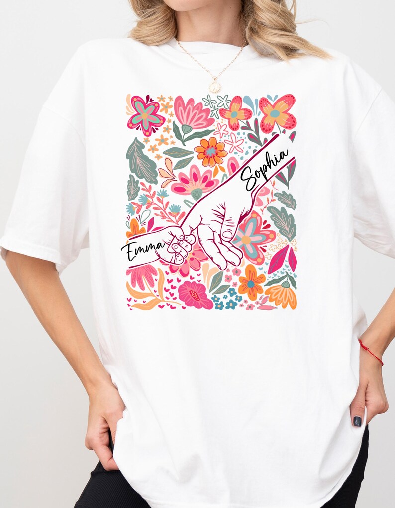 Op de afbeelding: Wit T-shirt met een bloemenontwerp met roze, oranje en blauwe bloemen. De afbeelding toont twee handen die elkaar vasthouden, met de namen "Emma" en "Sophia" erop. Een casual kledingstuk voor dagelijks gebruik.